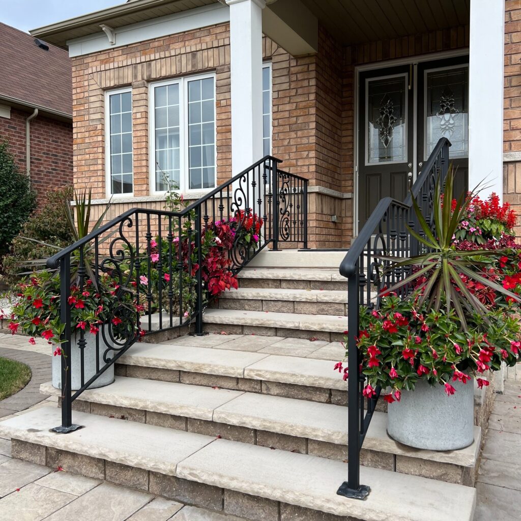 Custom fabricated iron railing with decorative scrollwork center feature installed on a concrete porch surrounded by flowers, crafted by Dream Ironwork with durable welded construction and elegant artistic detailing for enhanced curb appeal.